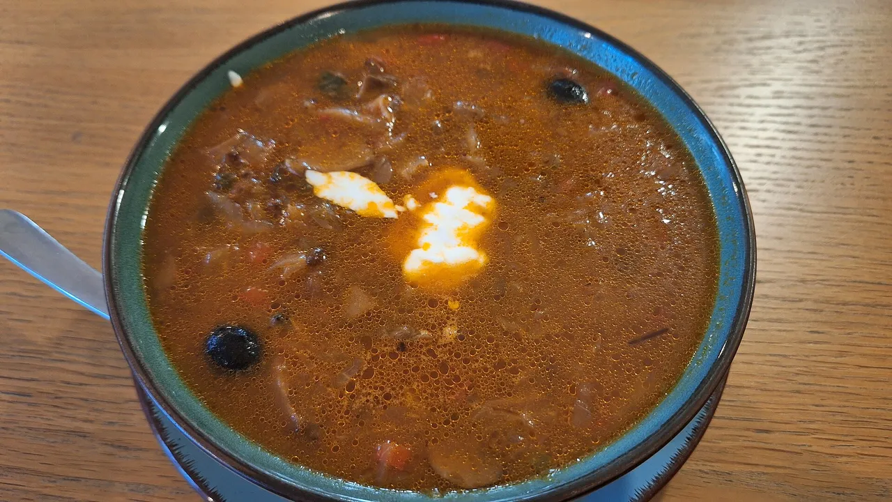 Мясная солянка со сметаной и маслинами в голубой глиняной миске на деревянном столе
